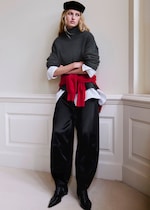 Placeholder for: Woman in a dark sweater, white shirt, red pullover tied at waist, wide black pants, and beret, leaning against a wall with crossed arms.