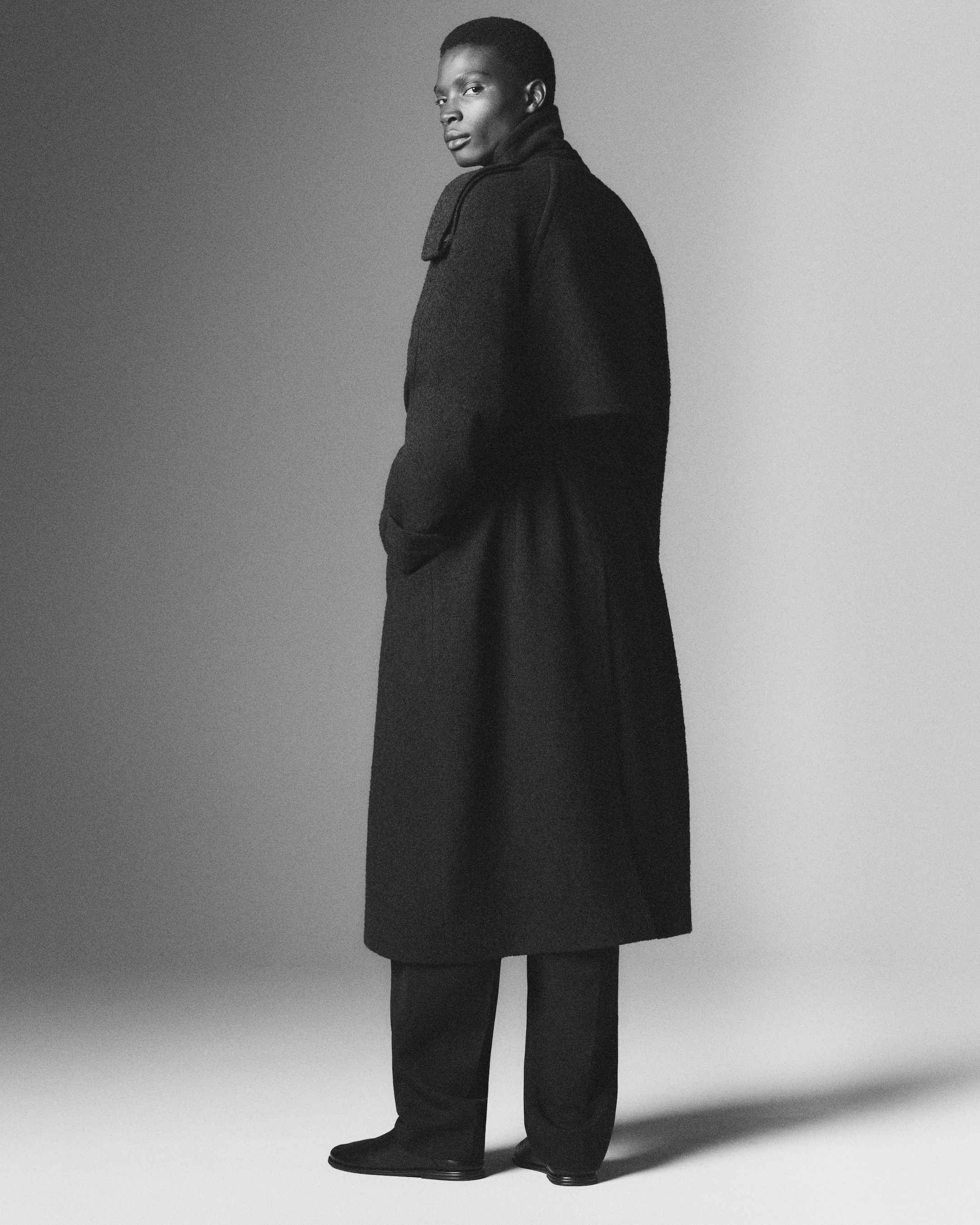 A person wearing a long, dark coat and trousers stands in a studio, slightly turning with a confident expression in a grayscale photograph.