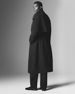 Placeholder for: A person wearing a long, dark coat and trousers stands in a studio, slightly turning with a confident expression in a grayscale photograph.