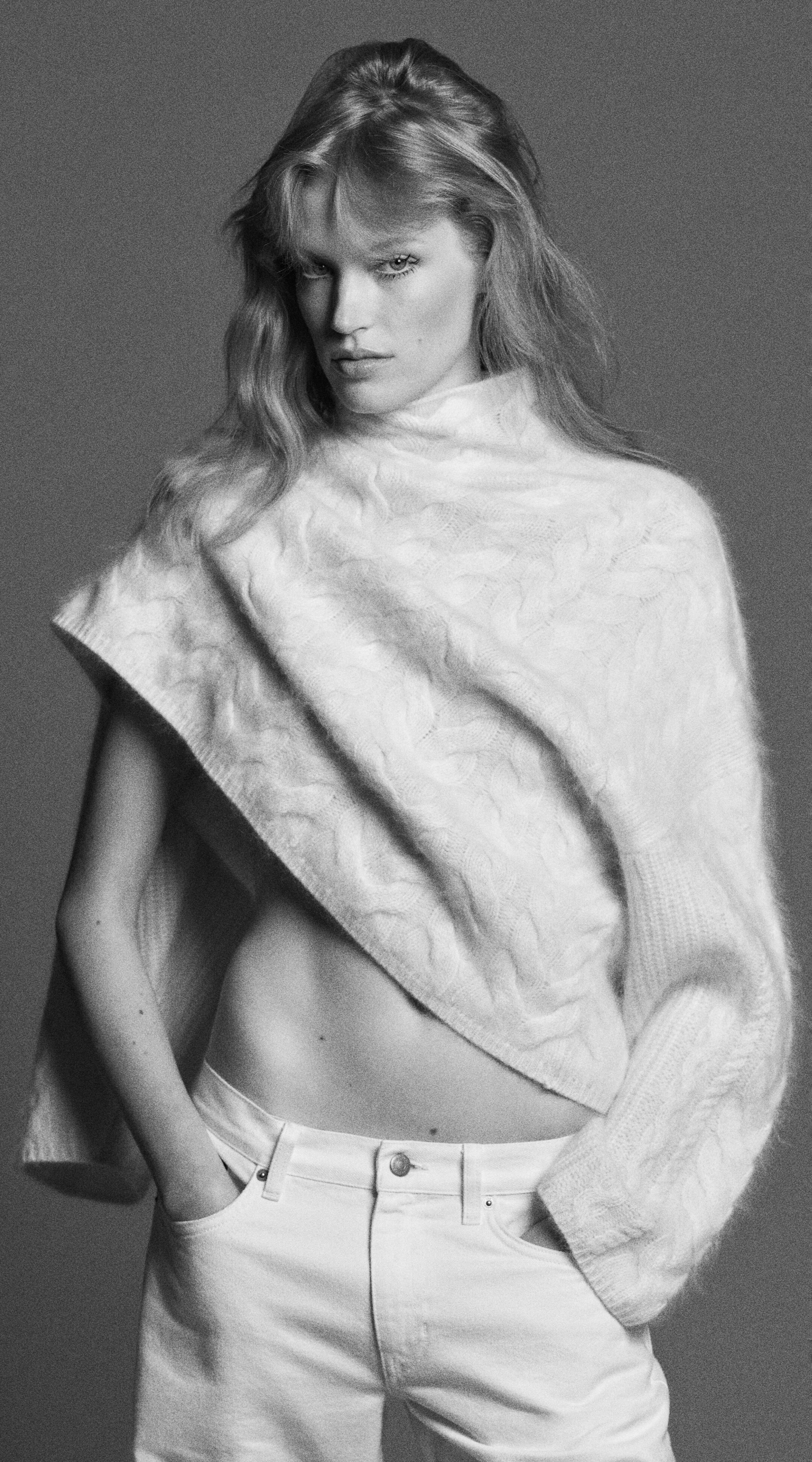 A person in a textured, draped sweater and white pants, with hands in pockets, gazing intensely in a black and white photo.