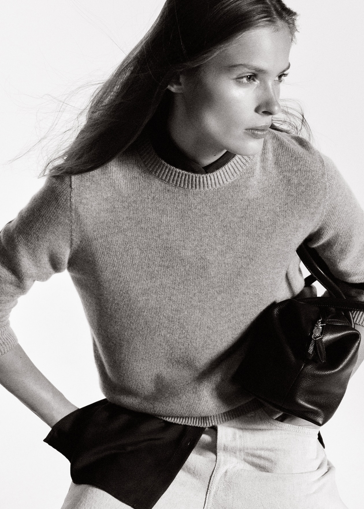 A black and white photo of a person with long hair, wearing a sweater and holding a small leather bag, looking to the side.