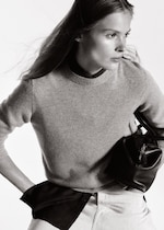 Placeholder for: A black and white photo of a person with long hair, wearing a sweater and holding a small leather bag, looking to the side.