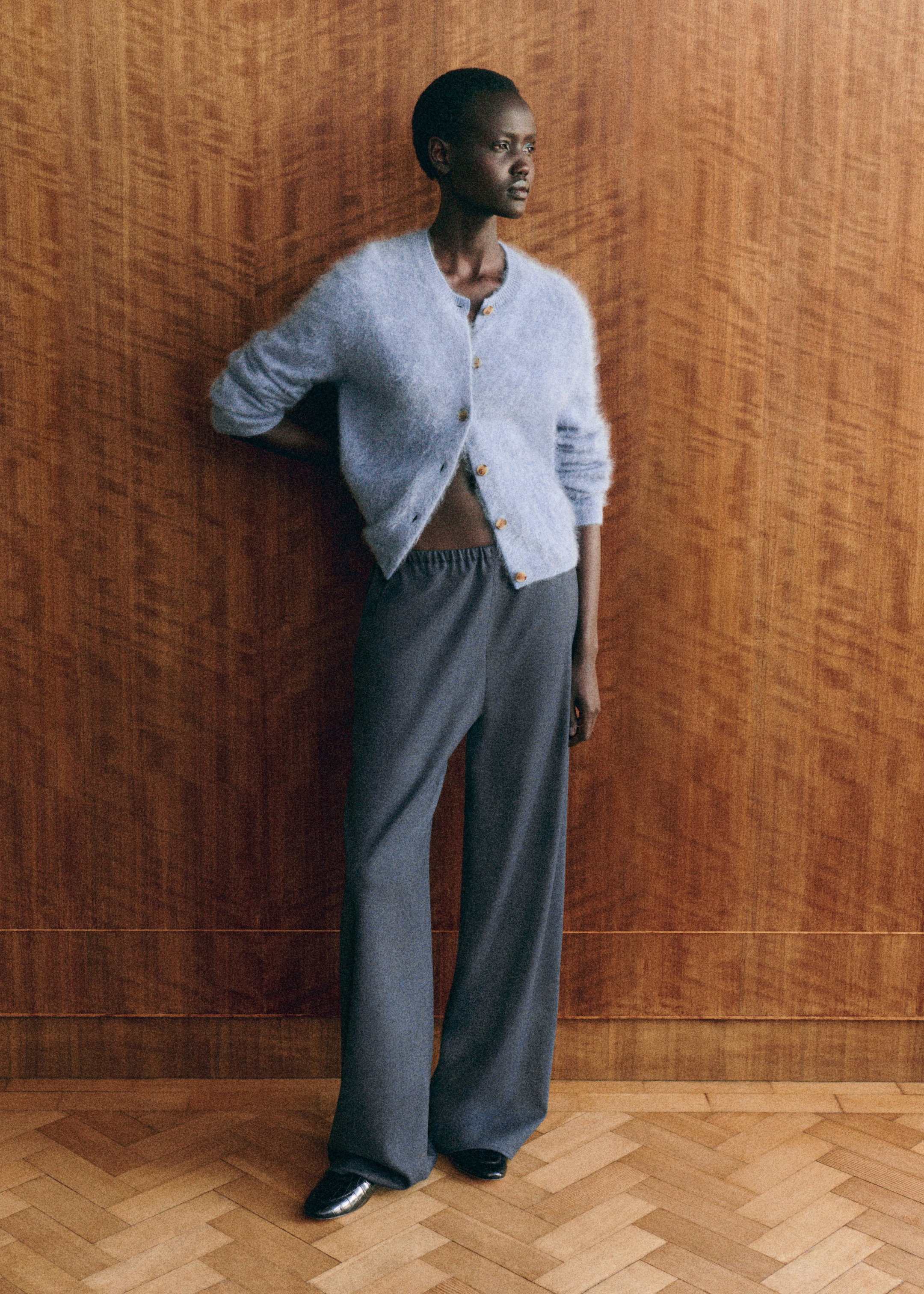A person in a fuzzy blue cardigan and gray pants stands against a wooden wall, gazing to the side.