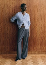 Placeholder for: A person in a fuzzy blue cardigan and gray pants stands against a wooden wall, gazing to the side.