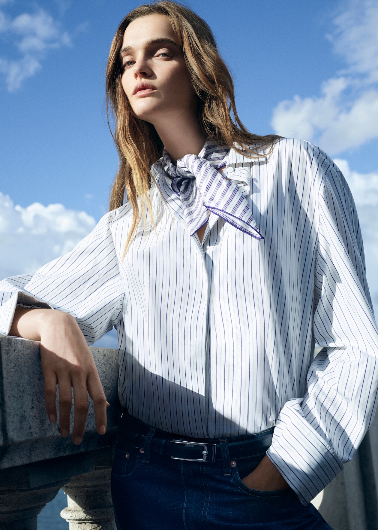 Person in a striped shirt with a neck scarf leans against a stone railing under a blue sky.