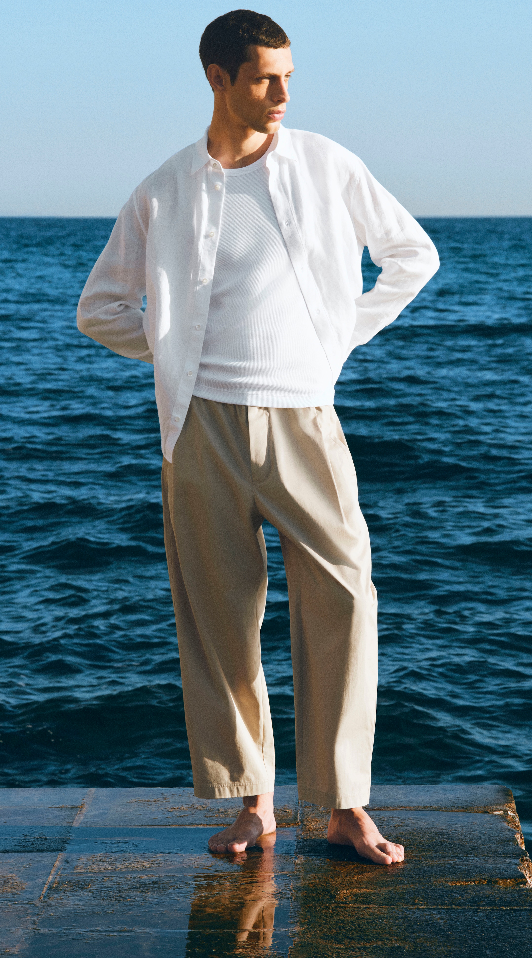Man in a white shirt and beige pants stands barefoot on a wet wooden surface by the ocean under a clear blue sky.