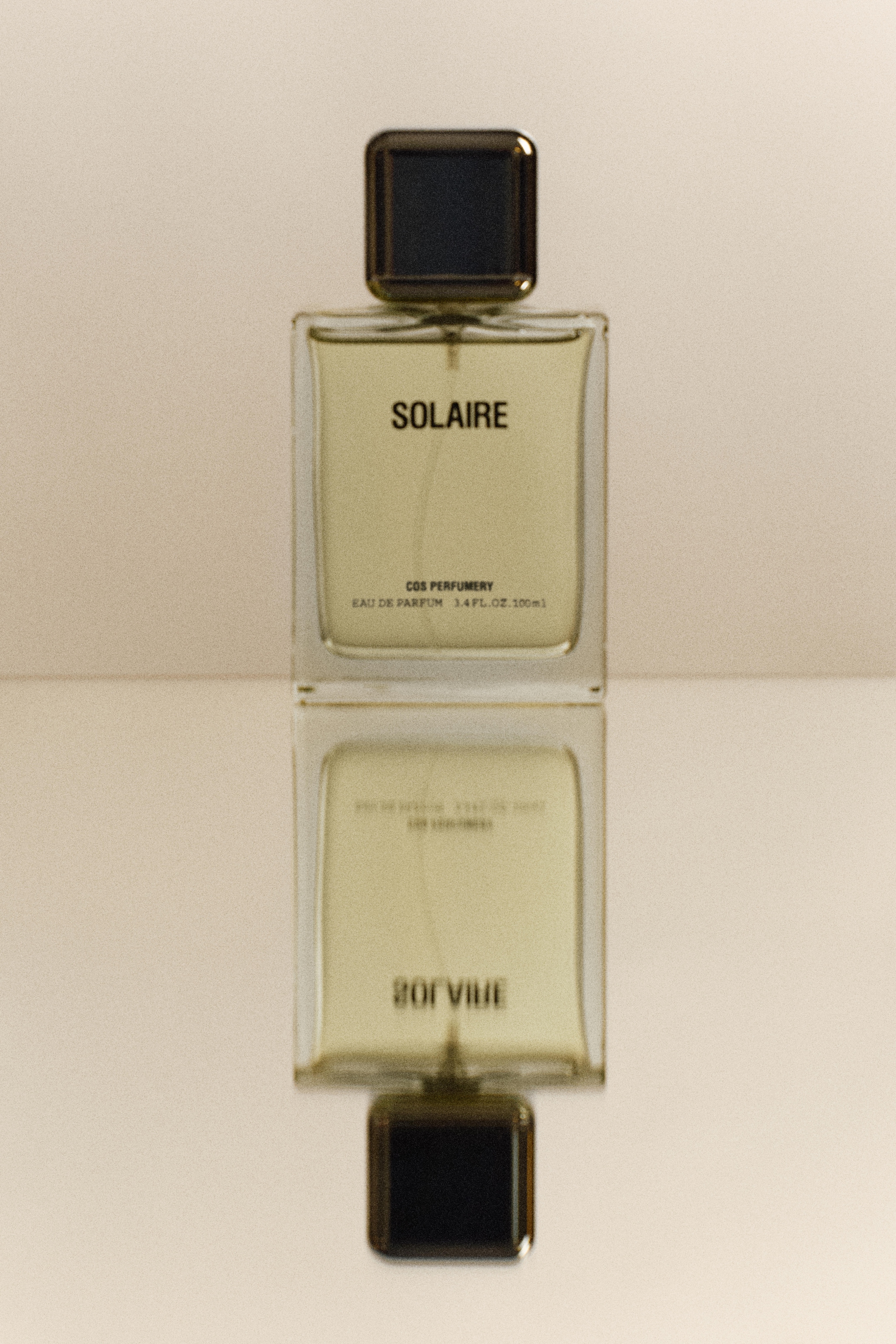 Perfume bottle labeled "Solaire" with dark cap, reflected on a glossy surface, creating a mirrored effect.
