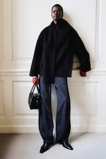 Placeholder for: A person stands against a wall, wearing a black coat, dark pants, and red gloves, holding a black handbag with a confident expression.