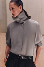 Placeholder for: Person wearing a high-collared, buttoned grey shirt with the sleeves rolled up, paired with black pants and a shiny black belt, in a neutral setting.