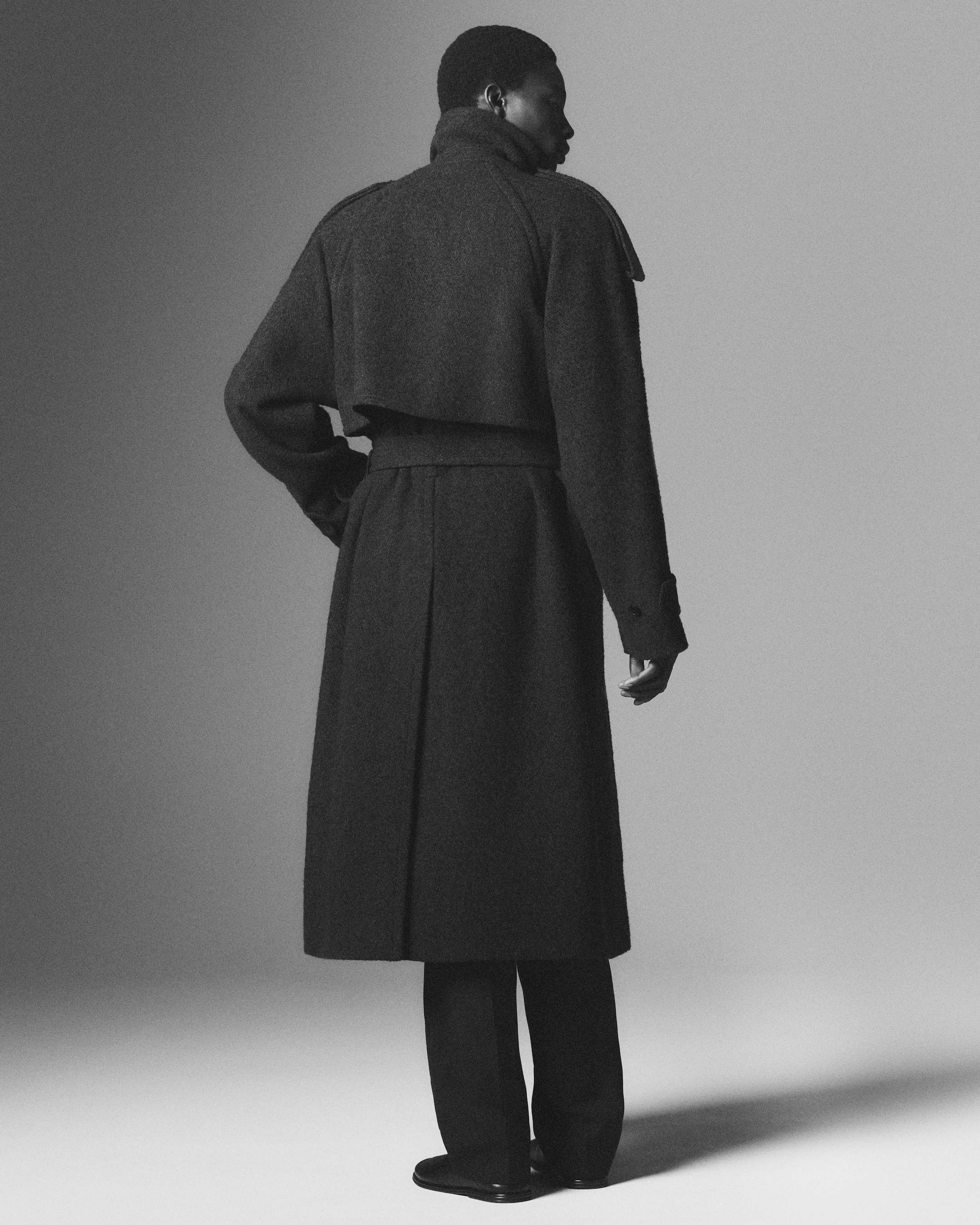 Person wearing a dark, long trench coat and trousers, standing with their back to the camera in a grayscale setting.