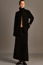 Placeholder for: A person wearing a long black buttoned outfit with a high collar poses against a plain background.