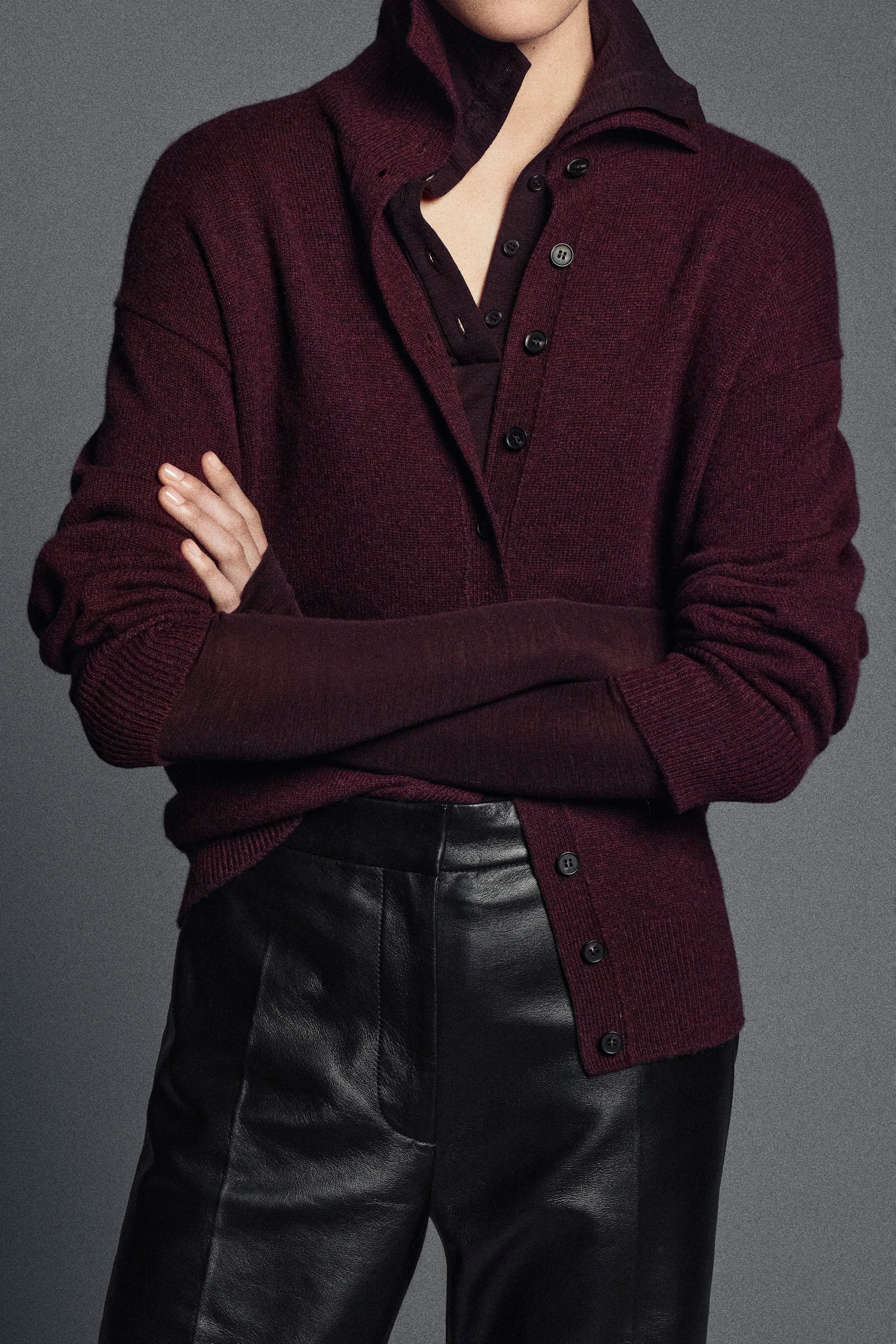 Person wearing a dark burgundy layered top, with sleeves rolled up, over black leather pants, standing with arms crossed.