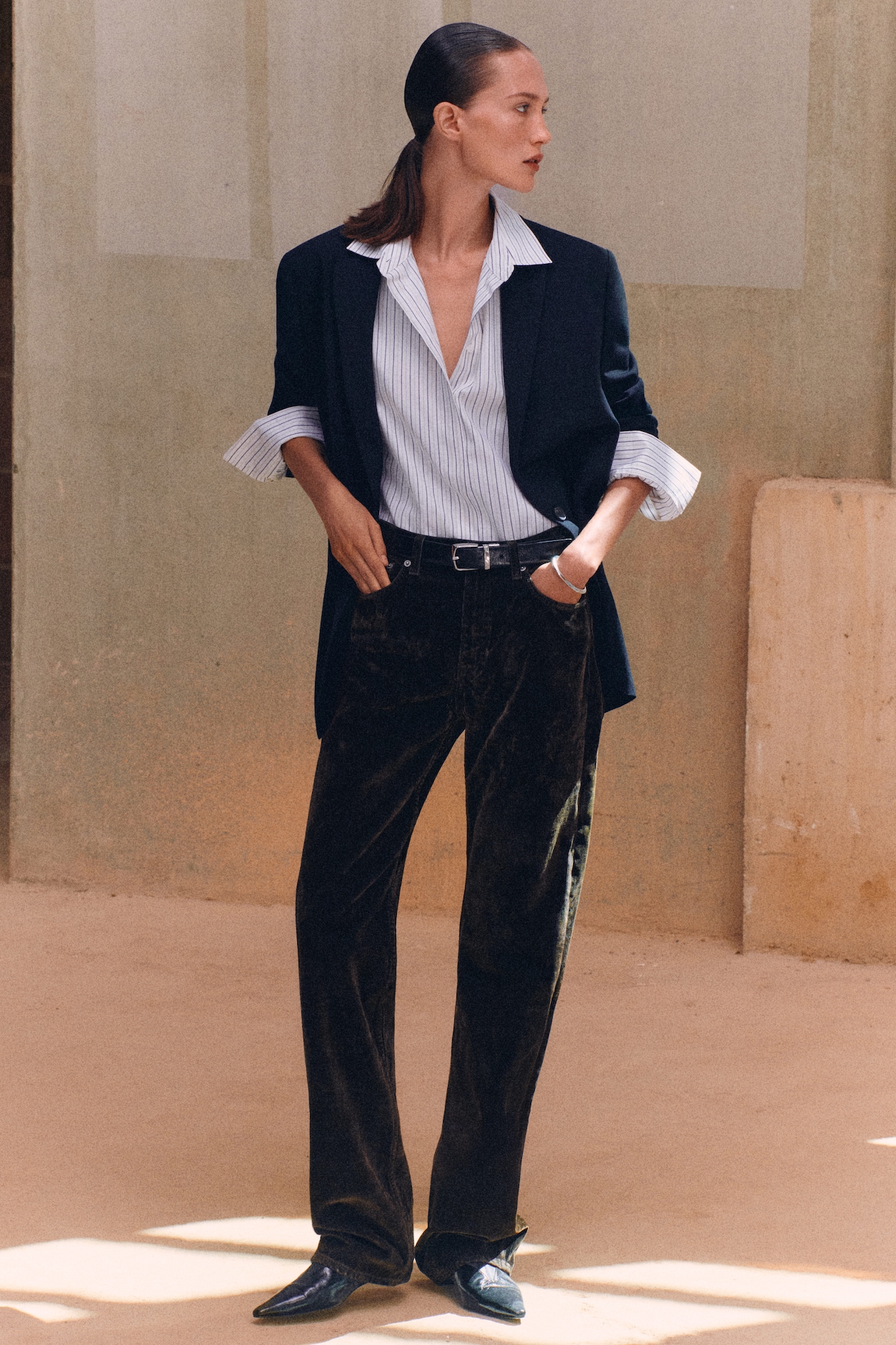 A person wearing a dark blazer, striped shirt, and dark trousers stands confidently in a minimalist setting.