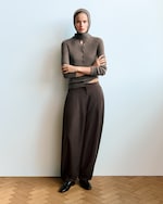 Placeholder for: A person wearing a gray sweater and headscarf stands with arms crossed, wearing loose brown pants and black shoes on a wooden floor.