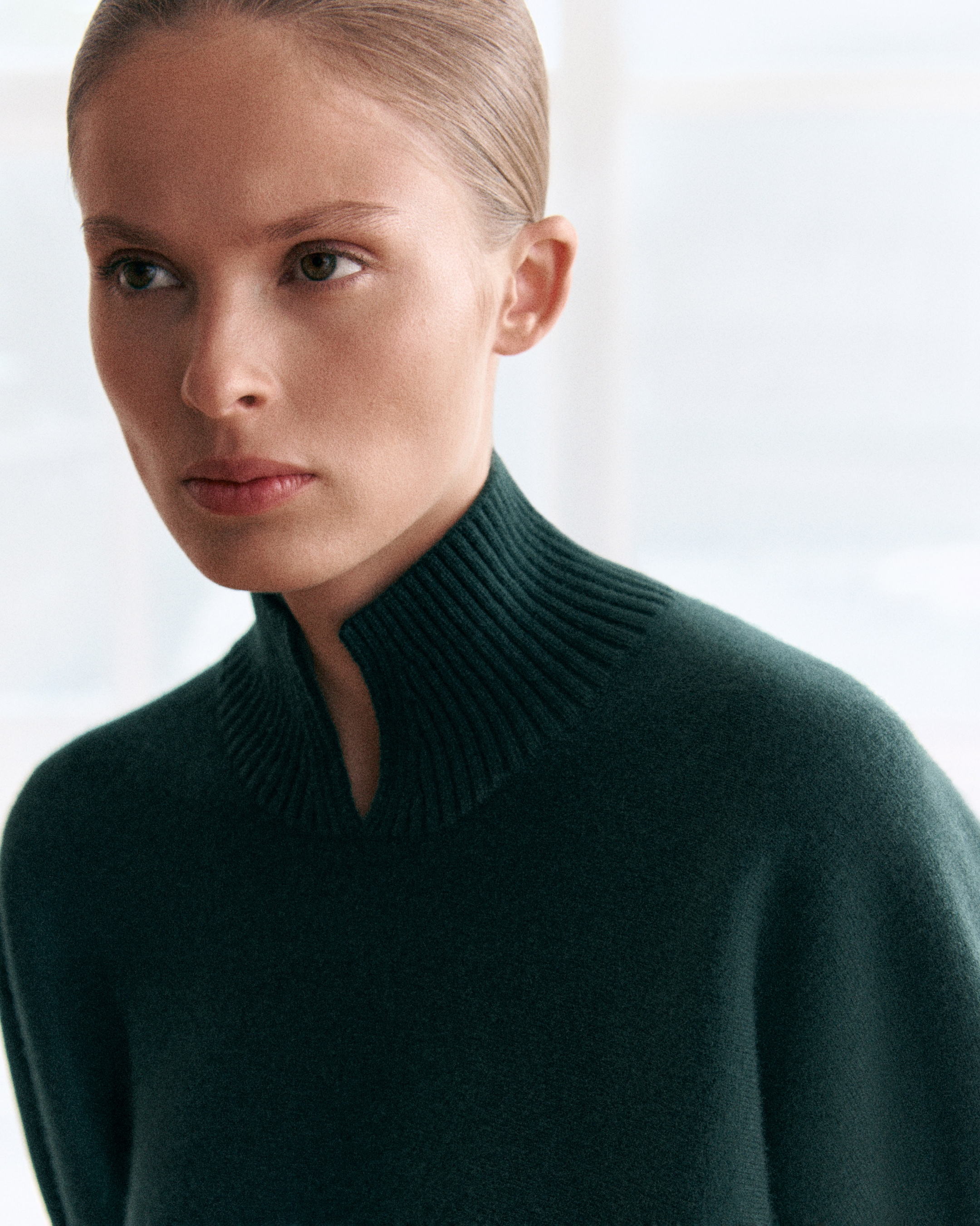 Person with light hair in a high-collared dark sweater, gazing thoughtfully to the side in a softly lit setting.