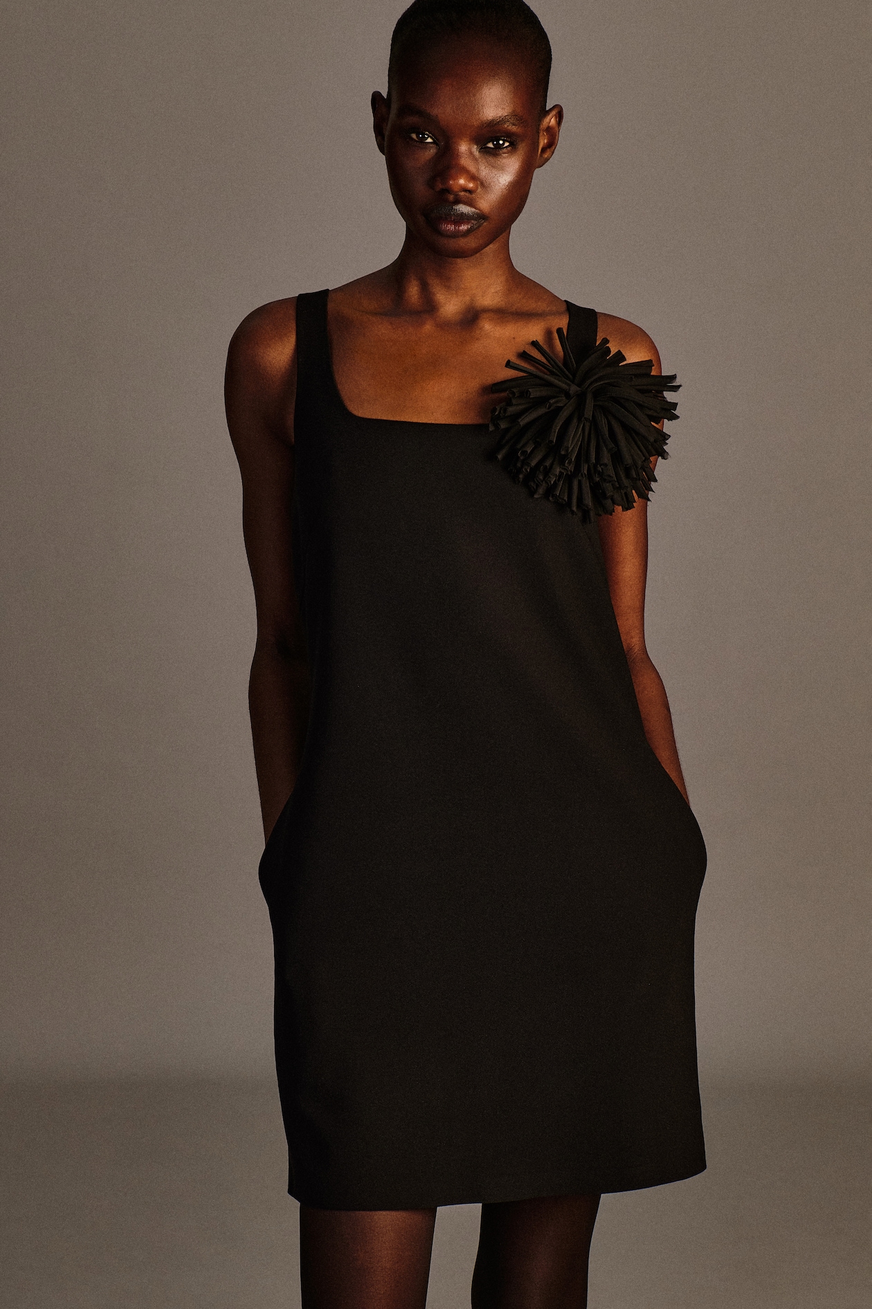 Person wearing a sleeveless black dress with a large floral embellishment on one shoulder, standing with hands in pockets against a neutral background.