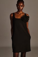 Placeholder for: Person wearing a sleeveless black dress with a large floral embellishment on one shoulder, standing with hands in pockets against a neutral background.