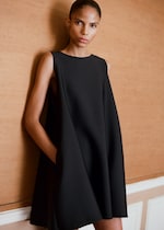 Placeholder for: A person with a sleek hairstyle wearing a black sleeveless dress, standing against a textured brown wall.