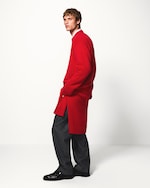 Placeholder for: Man in a red sweater and gray pants poses against a white background, looking to the side with hands in pockets.
