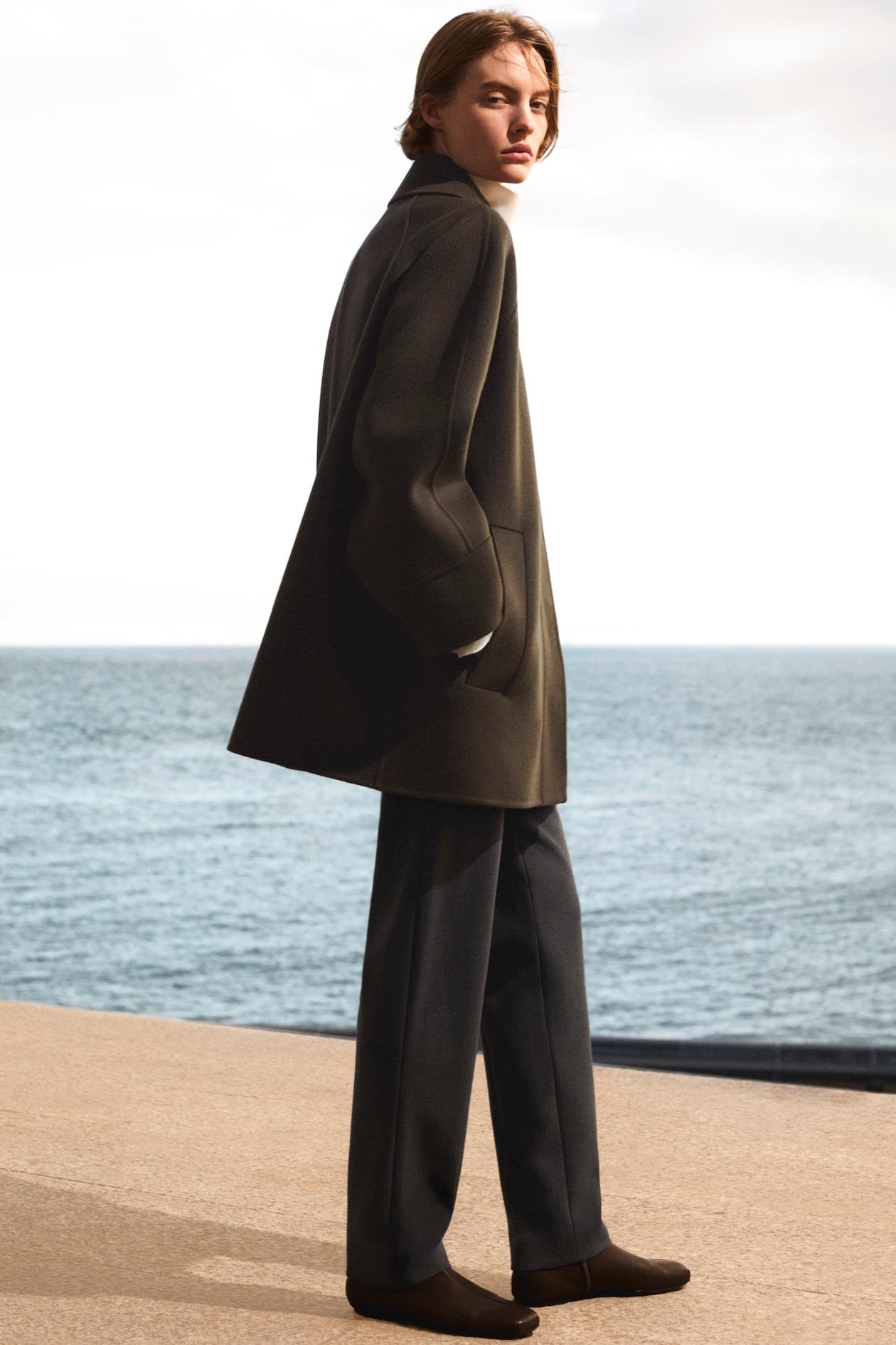 Person in an oversized dark coat stands on a concrete surface by the sea, with a cloudy sky in the background.