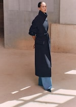 Placeholder for: Person standing indoors wearing a long black coat, blue jeans, and pointed shoes, with hands behind their back, casting shadows on the floor.