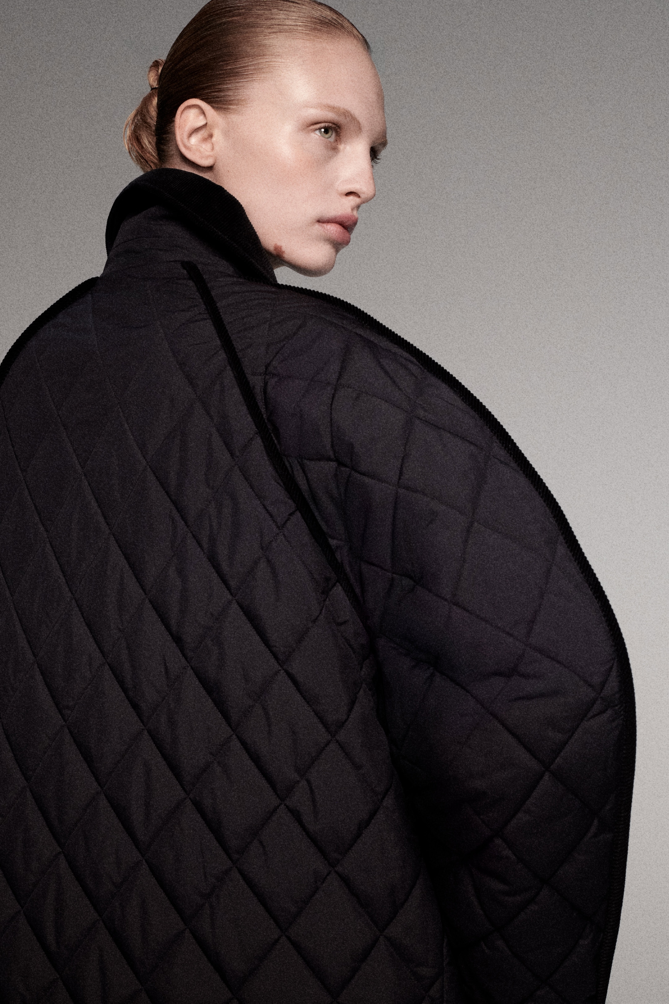 Person with blonde hair in a low bun, wearing a large black quilted jacket, looking to the left against a gray background.