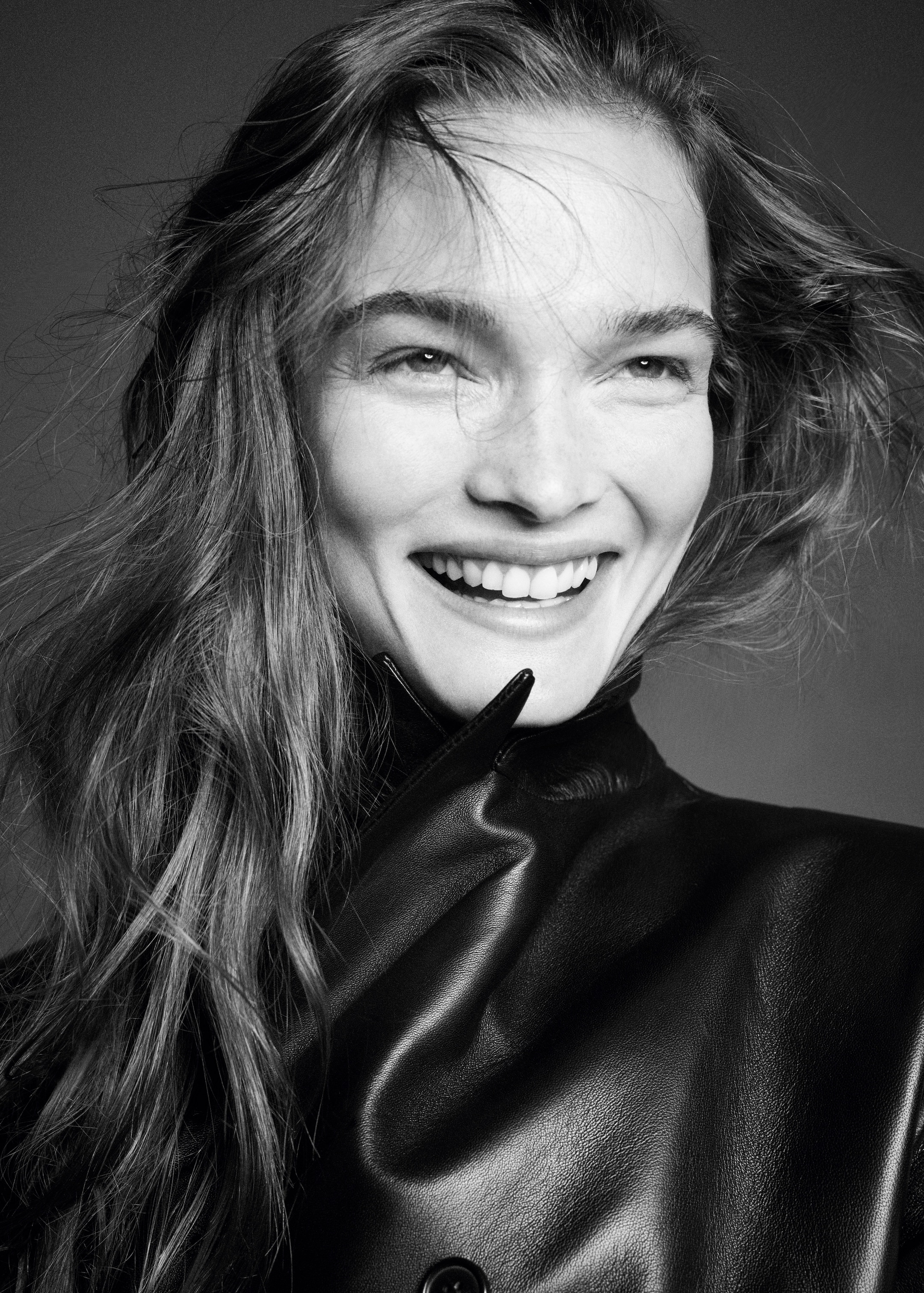 Black and white photo of a smiling person with loose hair, wearing a dark leather jacket.