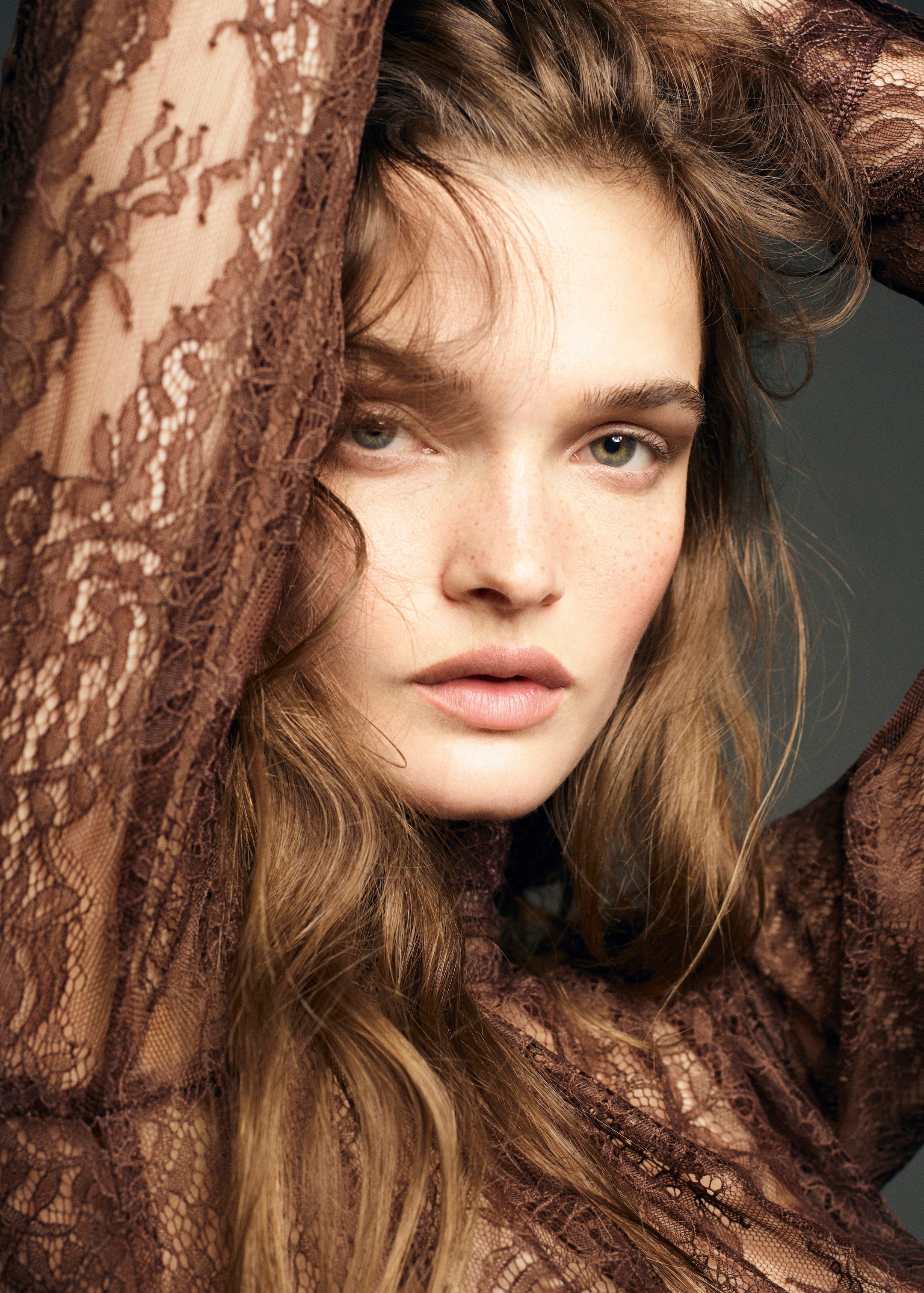 Woman with light brown lace top, wavy hair, arms raised, gazing directly at the camera against a muted background.