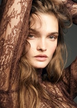 Placeholder for: Woman with light brown lace top, wavy hair, arms raised, gazing directly at the camera against a muted background.