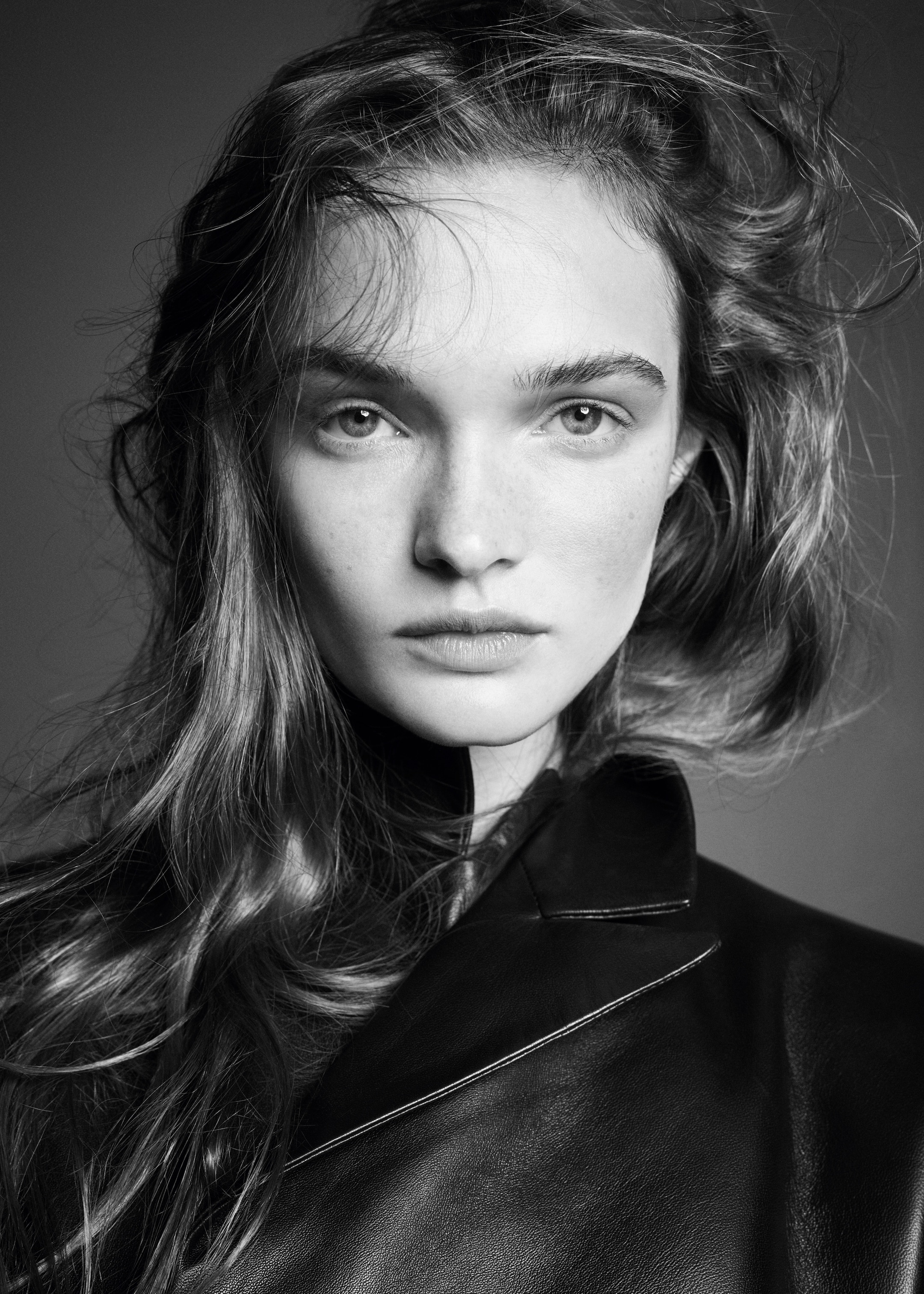 Black and white portrait of a person with wavy hair wearing a leather jacket, looking directly into the camera with a neutral expression.