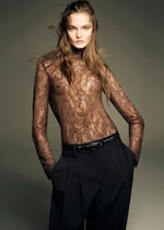 Placeholder for: A person wearing a sheer lace turtleneck top and black trousers with hands in pockets, posing confidently against a neutral background.