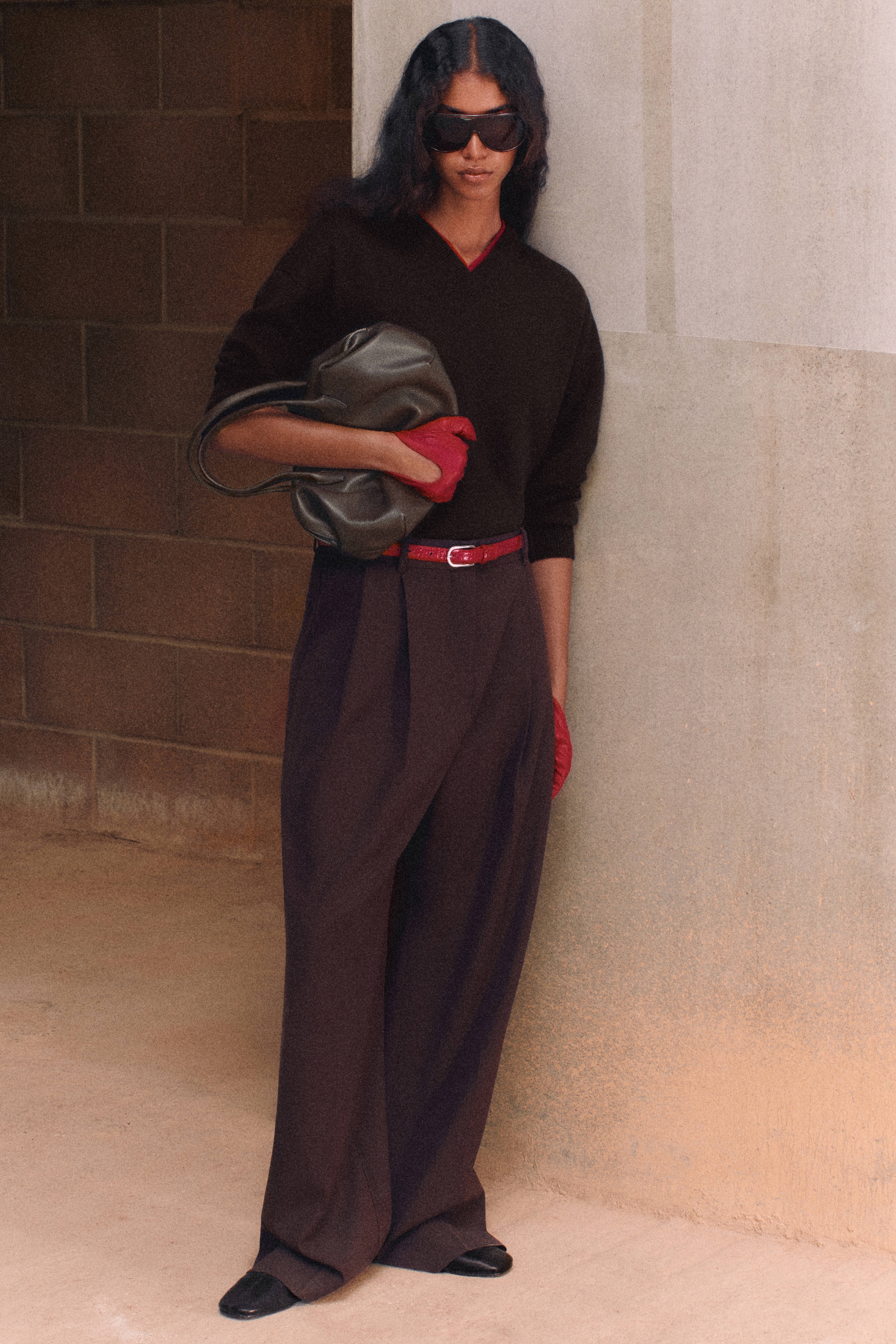 Person with long hair and sunglasses, wearing dark sweater, wide pants, red belt and gloves, holding a bag, leaning against a wall.