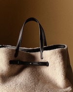 Placeholder for: A large, textured beige shearling tote bag with black leather handles and trim, set against a warm brown background.