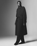 Placeholder for: A person in a long, dark coat with a high collar, standing in a minimalist setting, exuding a sense of elegance and poise.