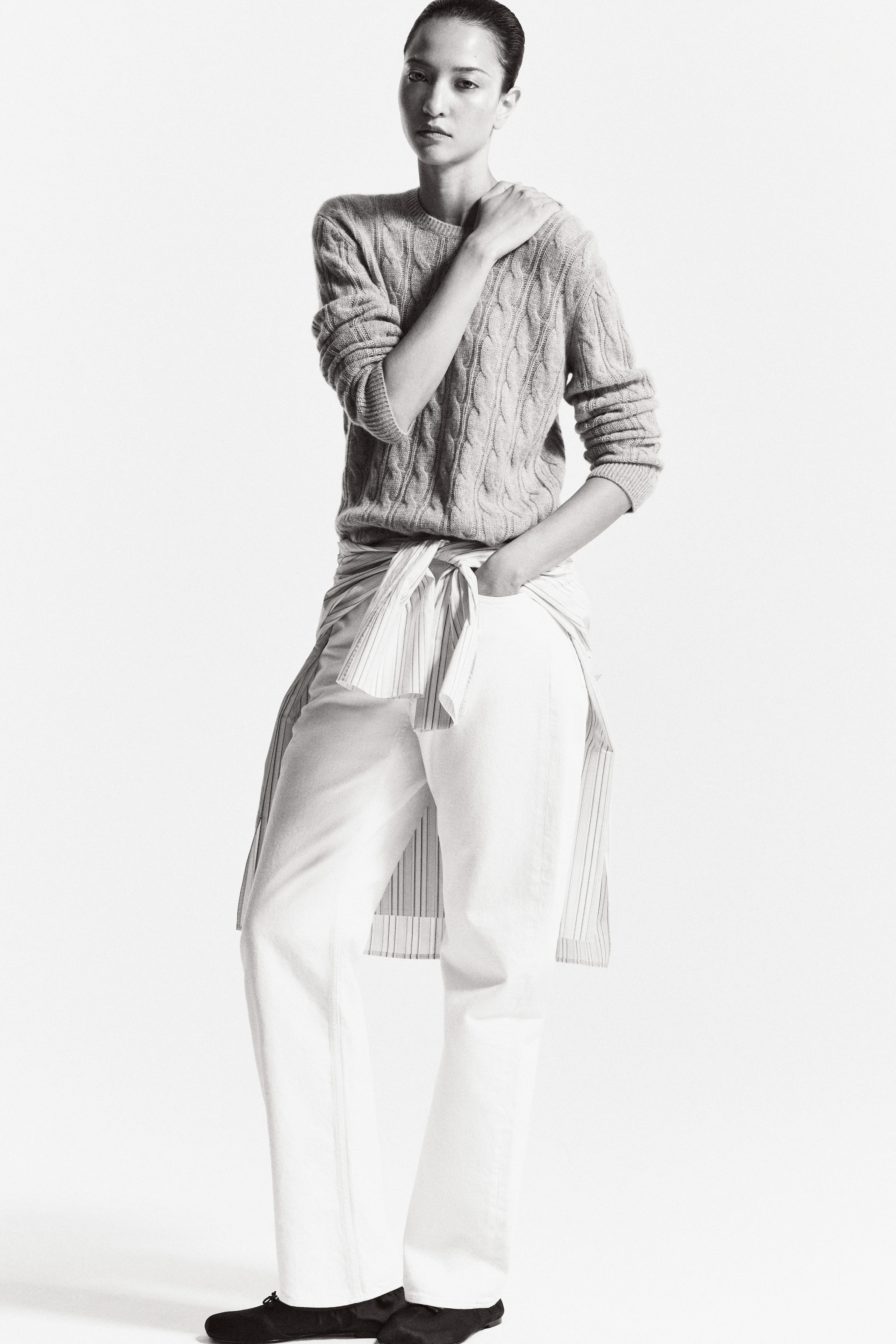 A person stands confidently wearing a textured sweater, white pants, and a striped shirt tied around the waist, with a neutral expression.