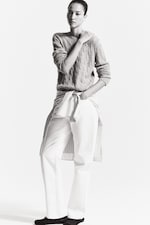 Placeholder for: A person stands confidently wearing a textured sweater, white pants, and a striped shirt tied around the waist, with a neutral expression.