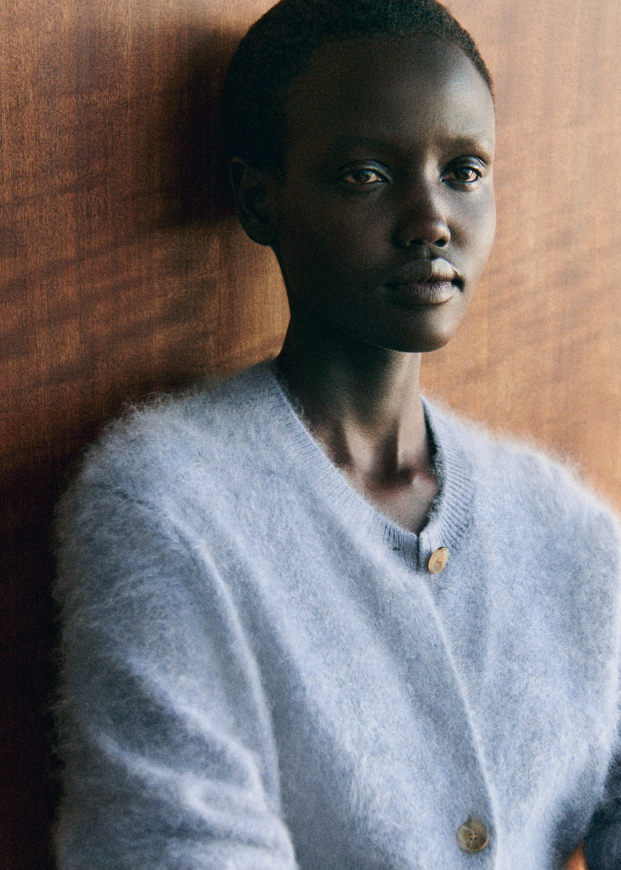 A person with short hair wearing a soft, light blue sweater leans against a wooden wall, looking thoughtfully into the distance.
