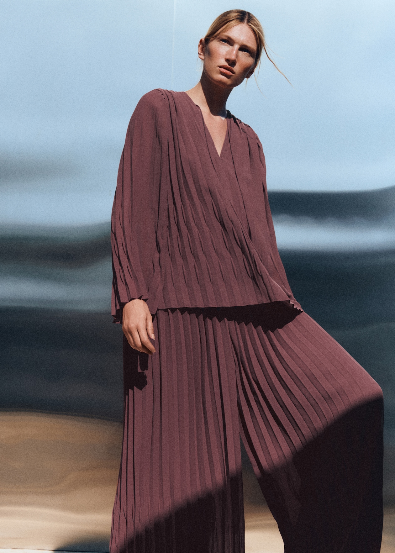 Person outdoors wearing a pleated mauve outfit, standing against a blurred background of earth tones and sky.