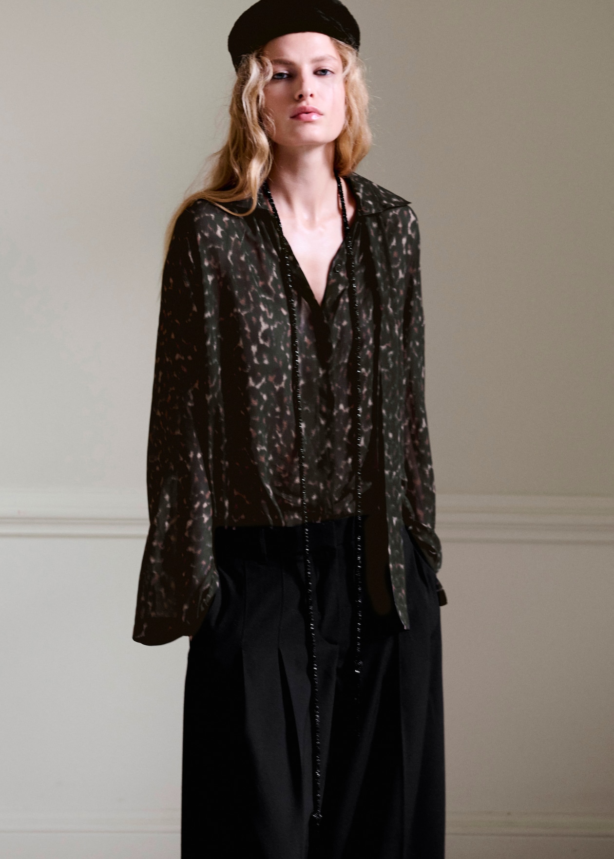 A person with long wavy hair wearing a black beret, a loose dark patterned blouse, and black trousers stands against a neutral background.