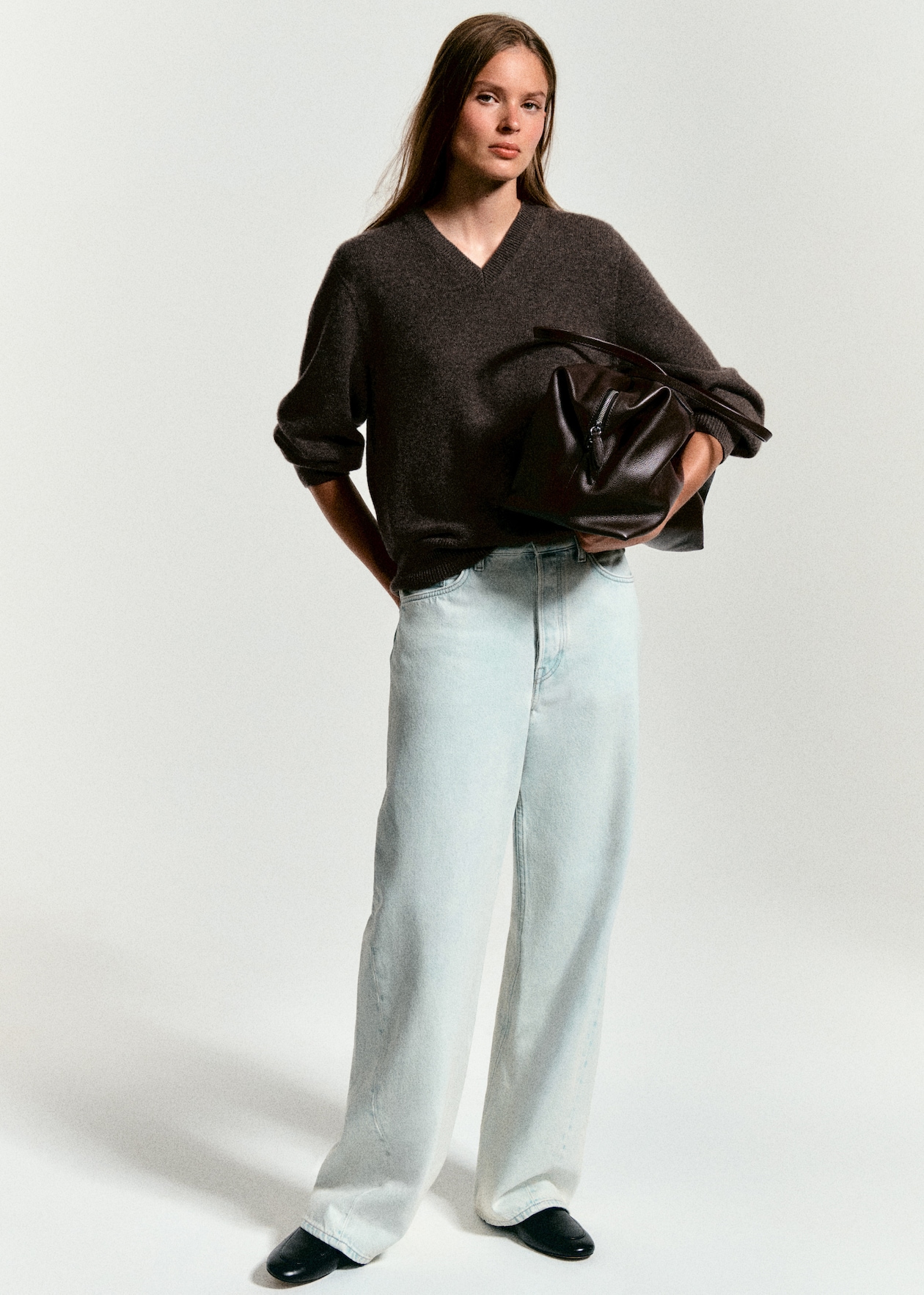 A person in a dark sweater and light jeans holds a large brown leather bag, standing against a plain background.