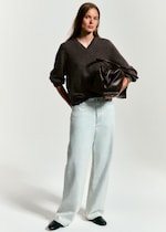 Placeholder for: A person in a dark sweater and light jeans holds a large brown leather bag, standing against a plain background.