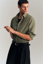 Placeholder for: Man in a green shirt and black pants, looking to the side with his hands clasped together, against a neutral background.