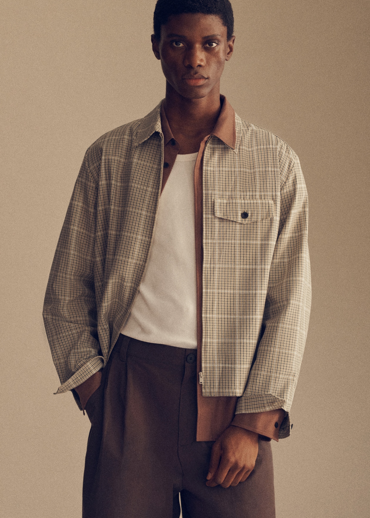 A person wearing a plaid jacket over a white shirt, with brown pants, stands against a neutral background.