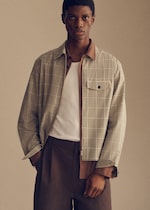 Placeholder for: A person wearing a plaid jacket over a white shirt, with brown pants, stands against a neutral background.