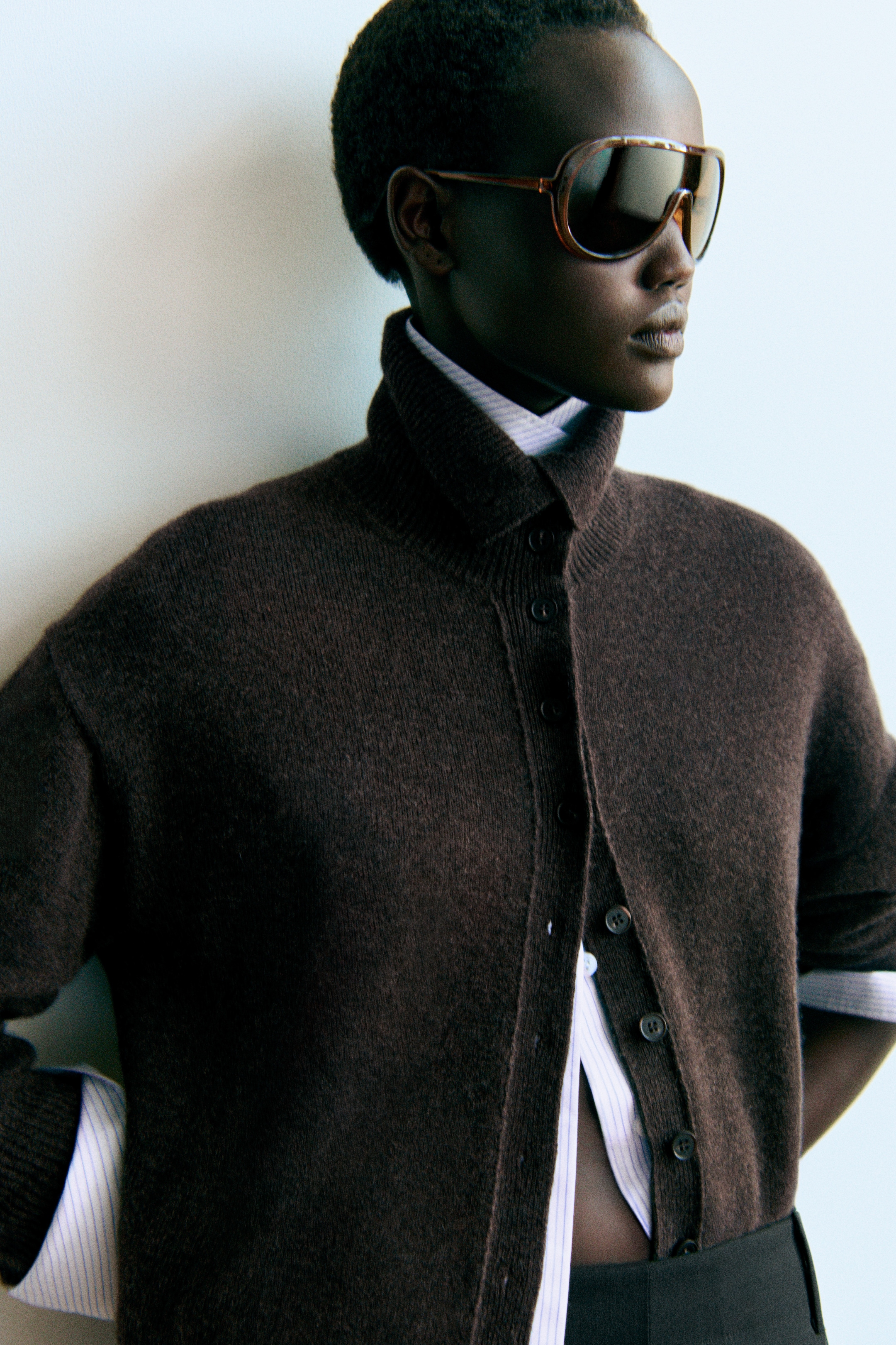 A person wearing oversized sunglasses and a brown buttoned sweater over a white collared shirt, standing against a light background.
