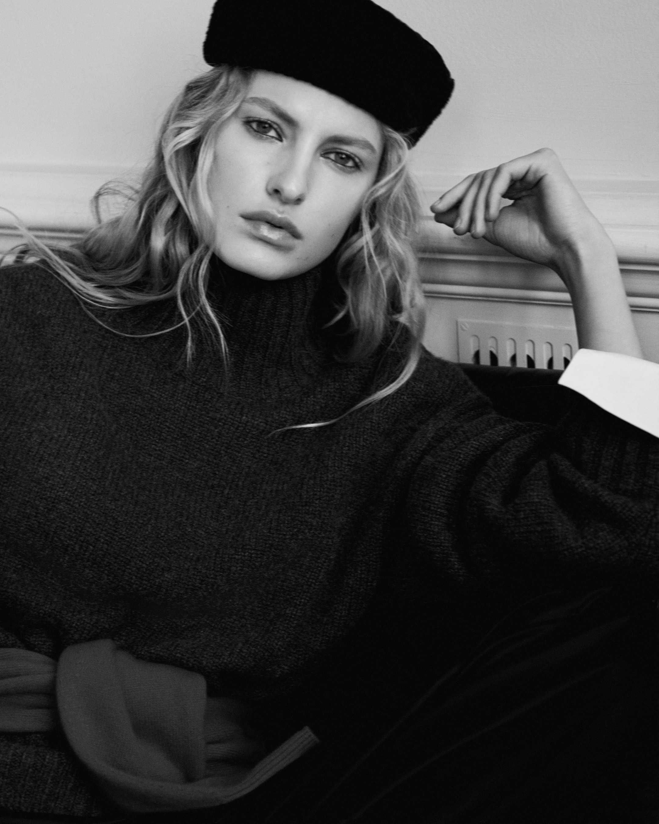 Black and white photo of a person wearing a dark beret and sweater, sitting with a pensive expression, hand resting on a knee.