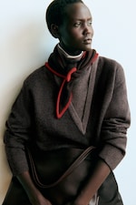 Placeholder for: A person in a brown sweater with a red scarf looks thoughtfully to the side, holding a large brown bag against a plain background.