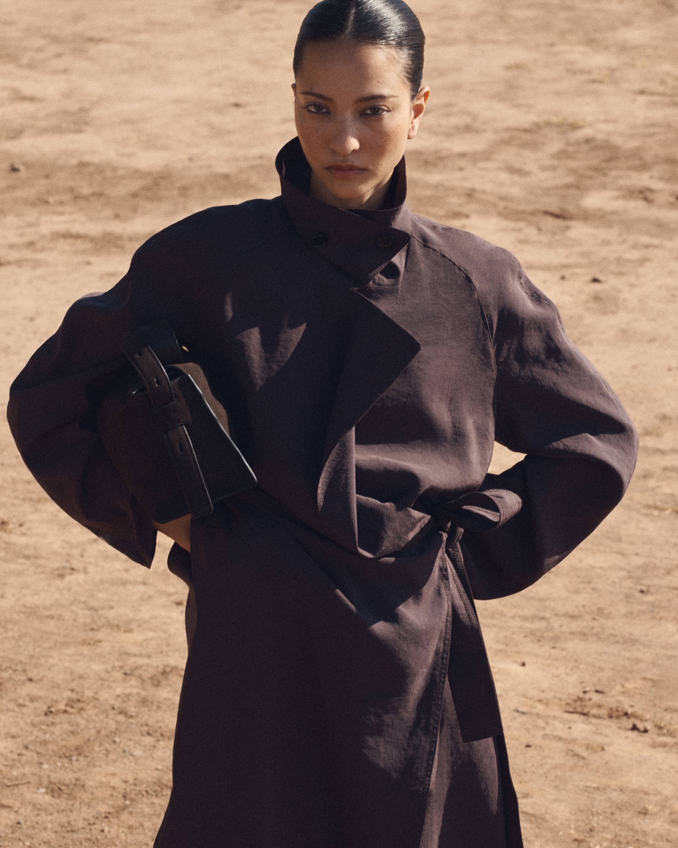 Person in a stylish dark brown coat stands on sandy terrain, holding a matching handbag, with a confident expression.
