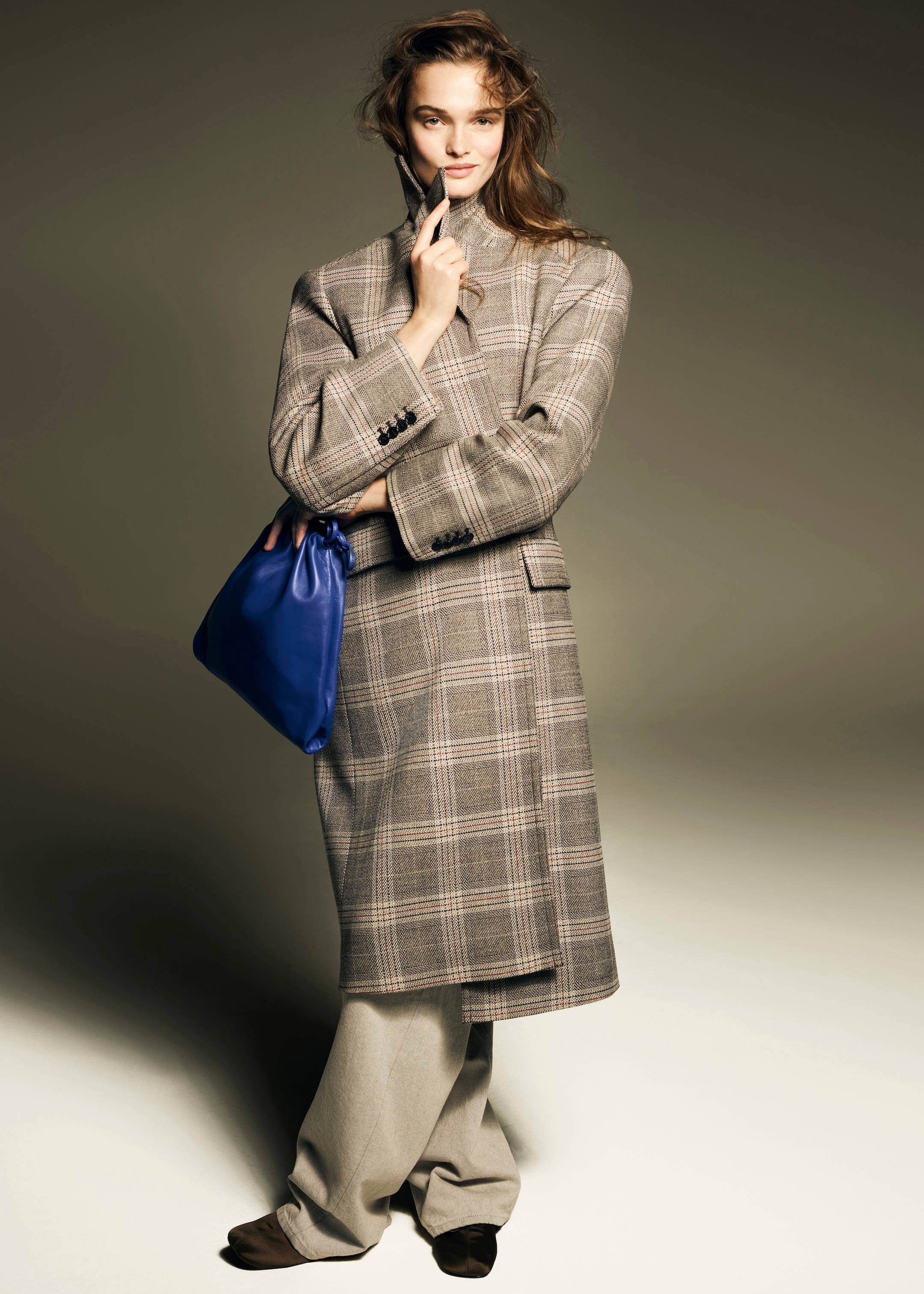 Woman in a plaid coat and wide trousers holds a blue bag. She stands against a neutral background, with a slight smile and tousled hair.