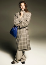 Placeholder for: Woman in a plaid coat and wide trousers holds a blue bag. She stands against a neutral background, with a slight smile and tousled hair.